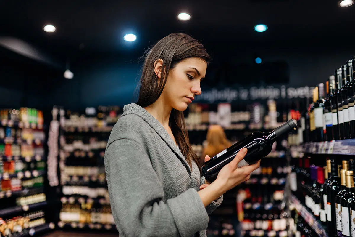 Woman examining a wine bottle in a store aisle, checking the label carefully.
