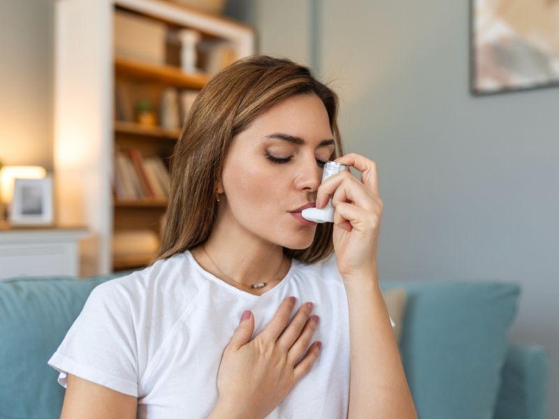 Woman with asthma using an inhaler while sitting at home, managing breathing difficulty effectively.