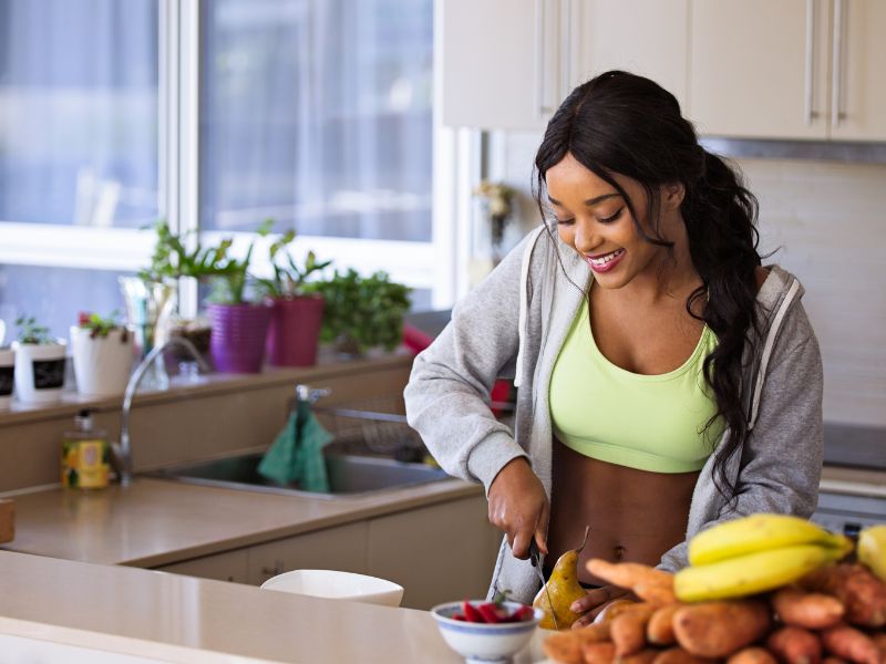 Smiling woman in a kitchen preparing fresh fruit, promoting a healthy lifestyle for improved sleep.