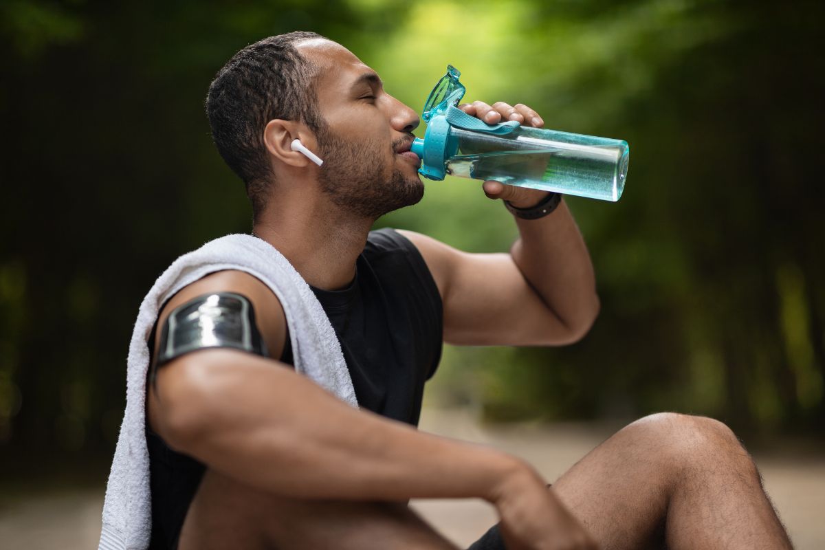 Man in workout attire drinking water from a bottle while resting outdoors, promoting healthy hydration.
