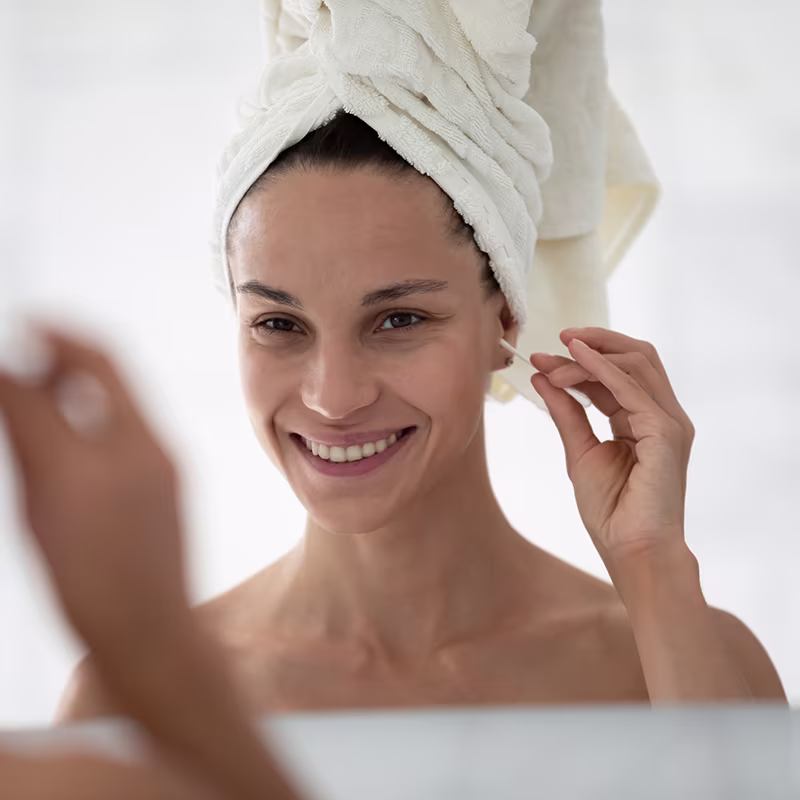 Smiling woman using cotton swab to clean ears, a common cause of ear wax impaction and ear canal irritation.