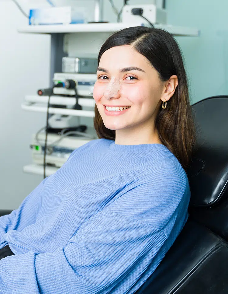 Smiling woman in clinic chair with nasal bandage, suggesting post-treatment recovery from a deviated septum.