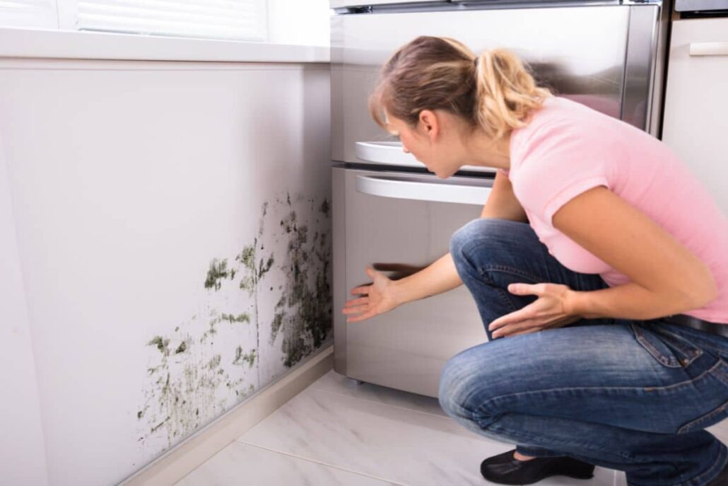 Concerned woman examining mold behind refrigerator in kitchen, showing health hazards of indoor mold buildup