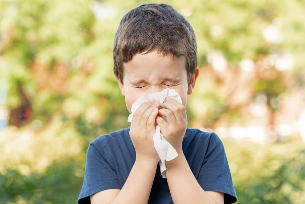 Young boy sneezing into tissue outdoors due to seasonal pollen allergies during summer.
