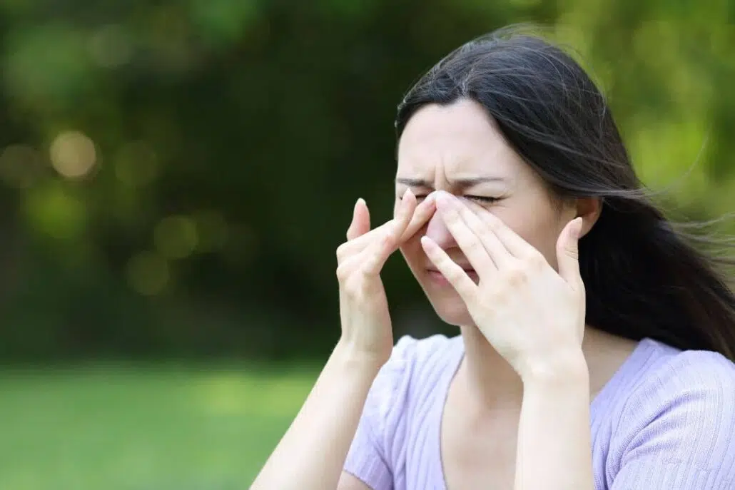 Woman rubbing eyes and nose outdoors, showing signs of tree pollen allergy like itchy eyes and sinus pressure.