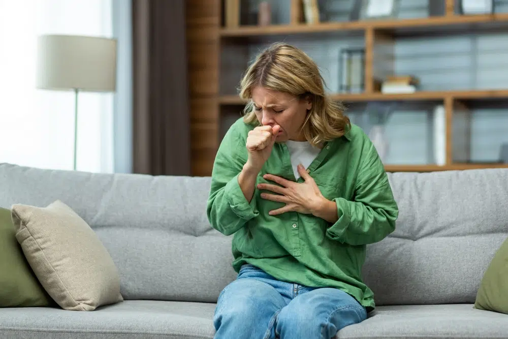 Woman coughing on sofa, holding chest, possibly experiencing swollen lymph nodes due to seasonal allergies.