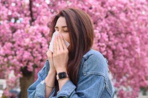 Woman sneezing due to spring allergies in Robbinsville, NJ with blooming pink trees in the background.