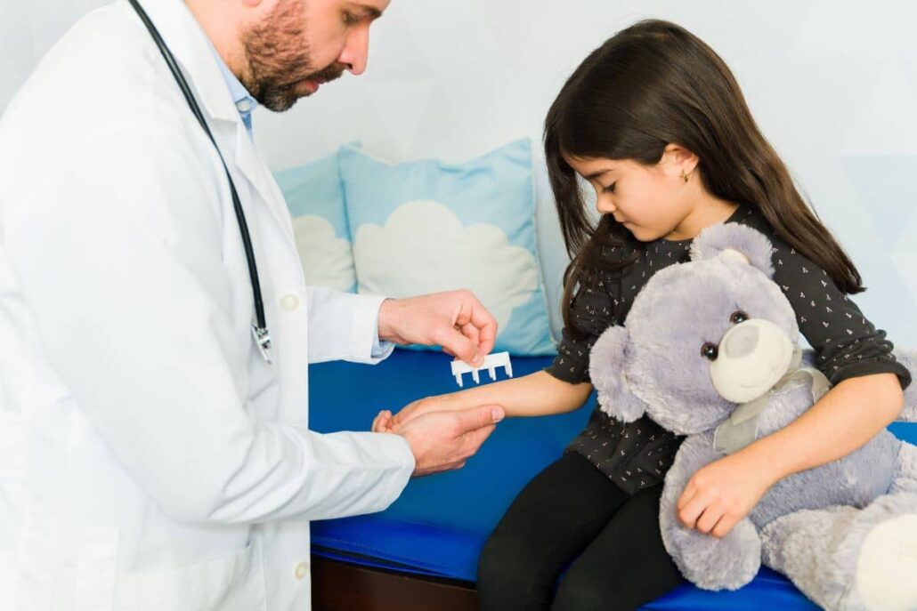 Doctor performing skin prick allergy test on young girl’s forearm in child-friendly clinic setting with stuffed animal.