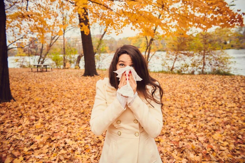 Woman outdoors in autumn covering her nose with a tissue, illustrating fall ragweed allergies and sinus pressure.