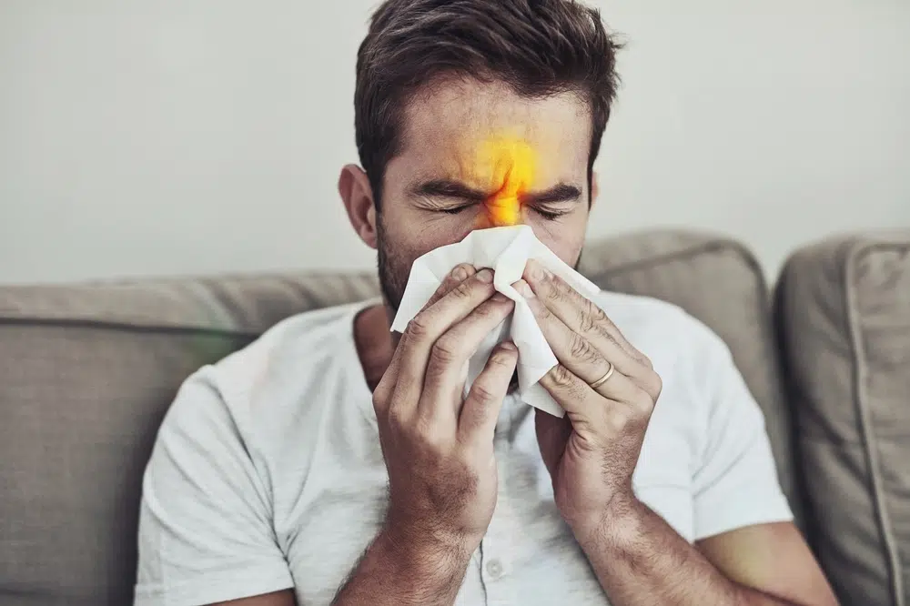 Man blowing nose with tissue while experiencing sinus pain and nasal congestion, highlighting inflamed sinus area on forehead.