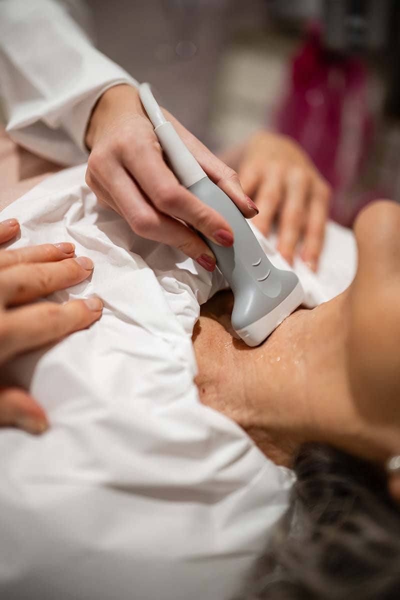 Doctor examining a patient's neck during a consultation for salivary gland evaluation, with diagnostic imaging equipment in the background.