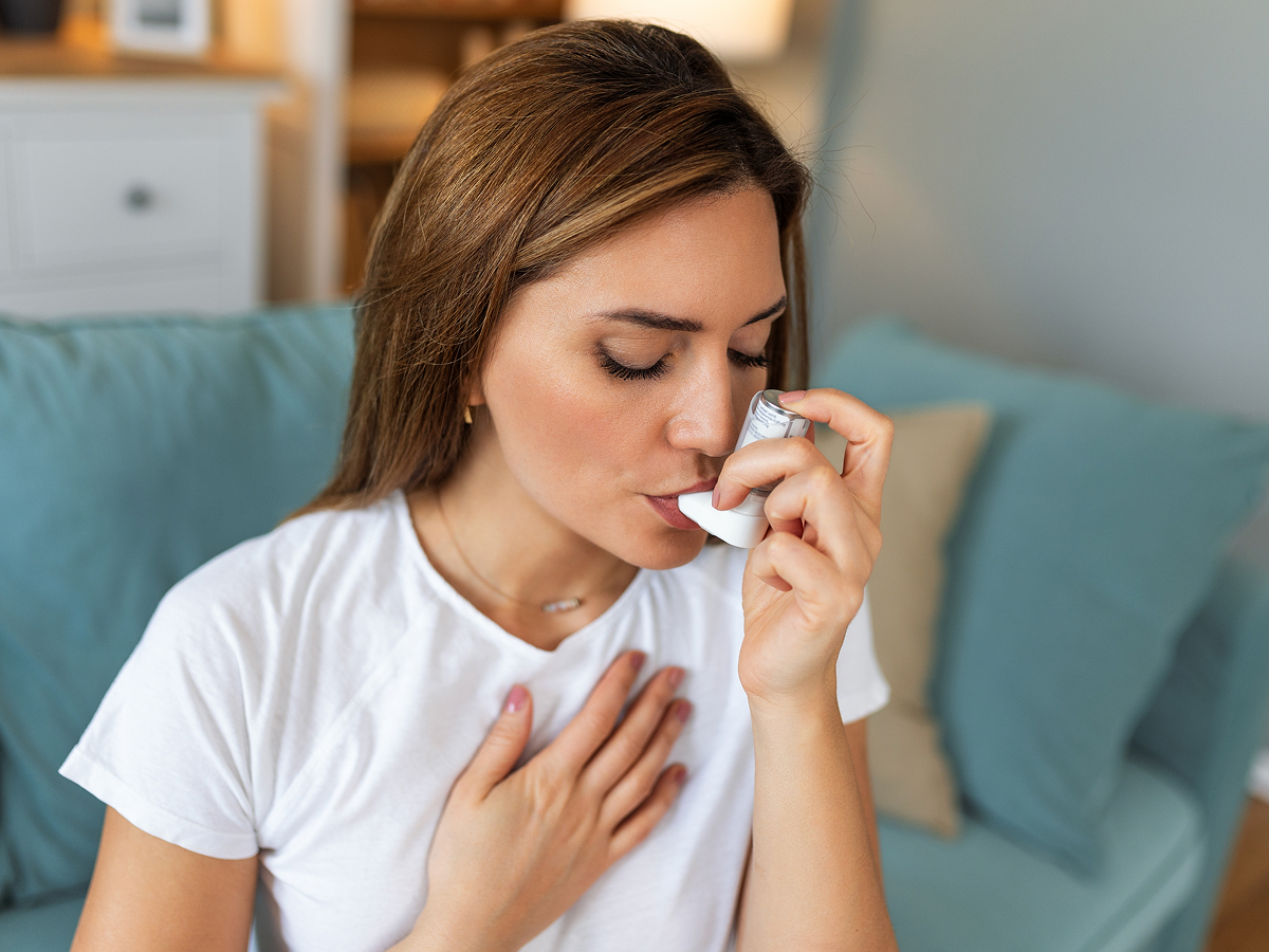 Woman using an inhaler at home for asthma treatment and symptom relief related to allergy triggers.