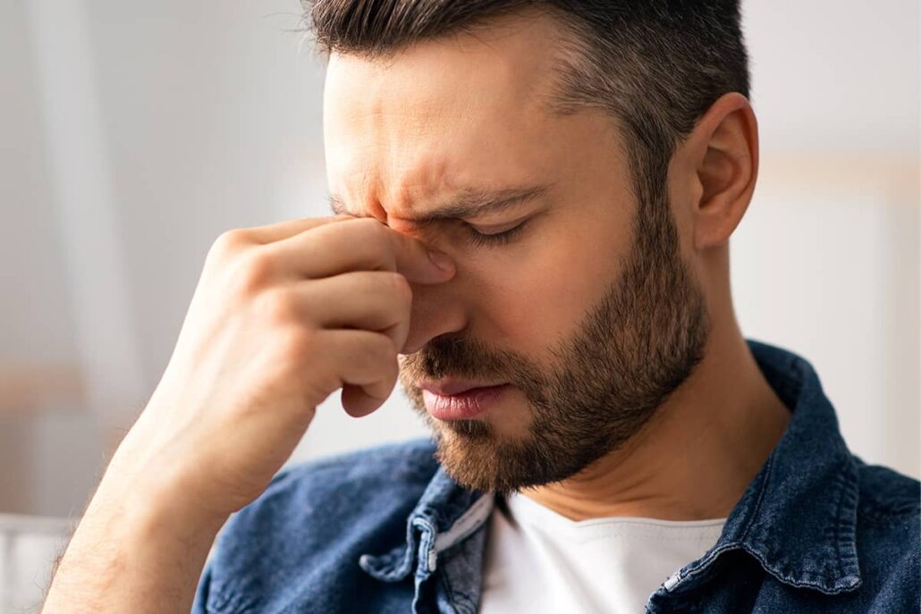 A close-up of a bearded man in a denim shirt closing his eyes in discomfort while pinching the bridge of his nose.