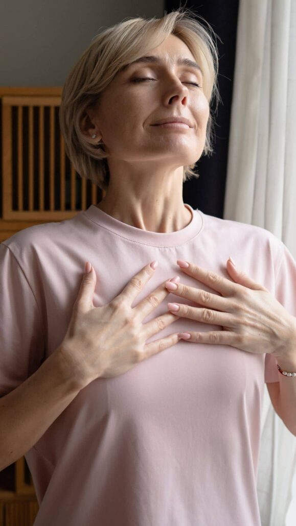 A blonde woman closing her eyes with a peaceful expression and resting her hands on her chest as she takes a deep, comfortable breath.