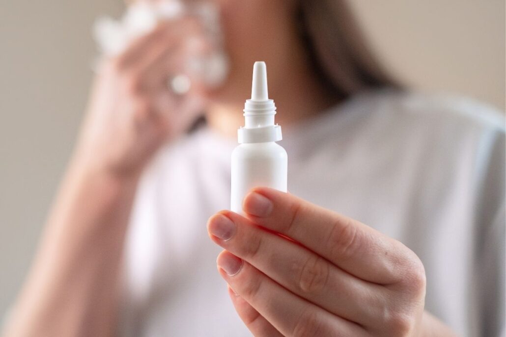 A close-up focus on a small white nasal spray bottle held in a hand, with a blurred person using a tissue in the background.