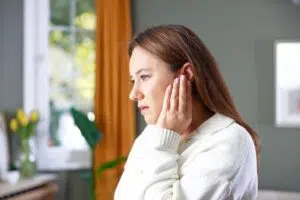 Woman holding her ear in discomfort while experiencing sudden ear pain at home near a window.