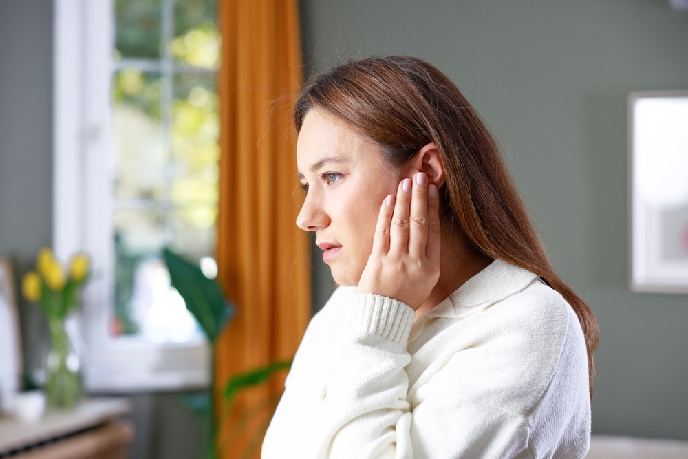 Woman holding her ear in discomfort while experiencing sudden ear pain at home near a window.